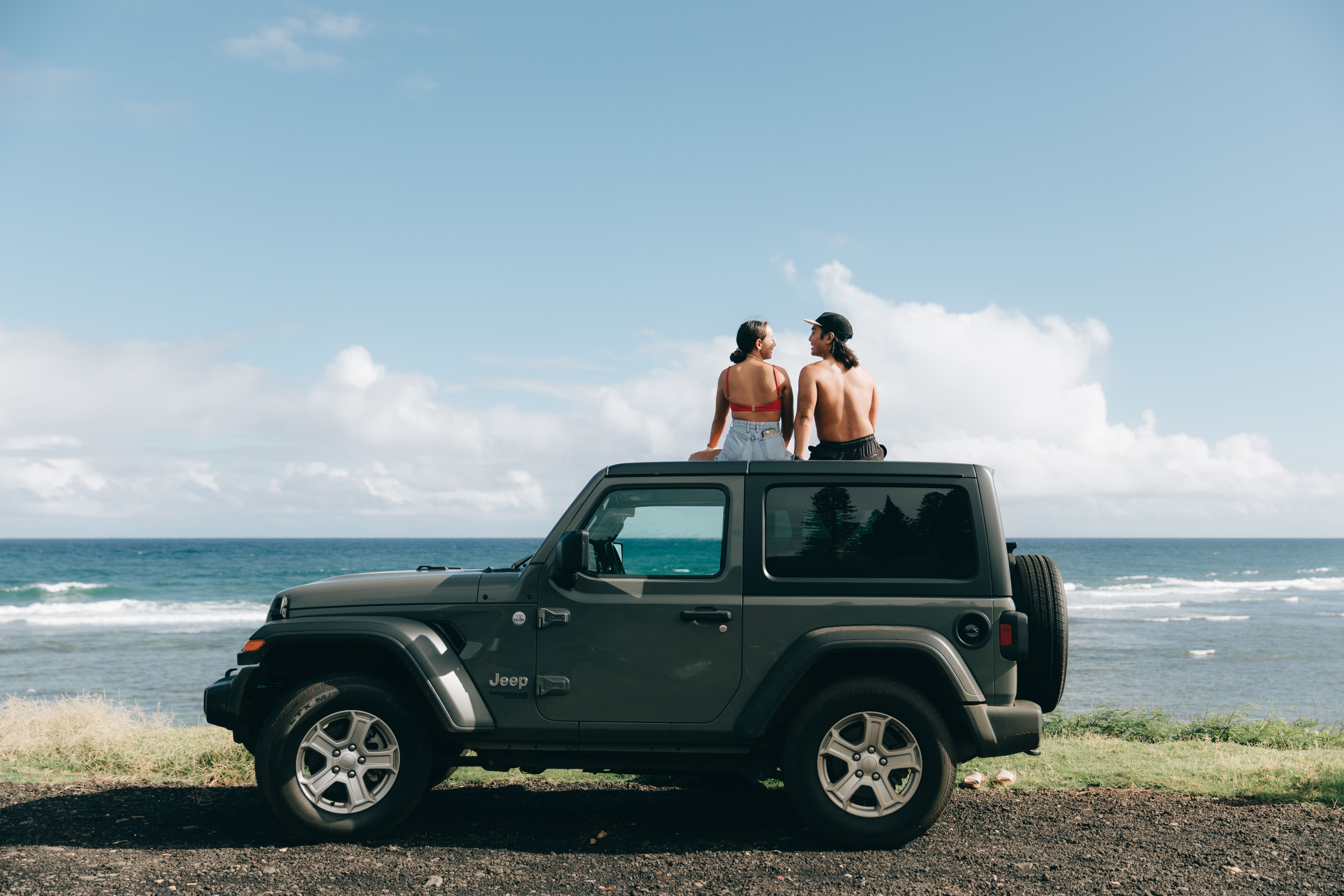 Two people sit on a Jeep by the ocean under a blue Honolulu sky
