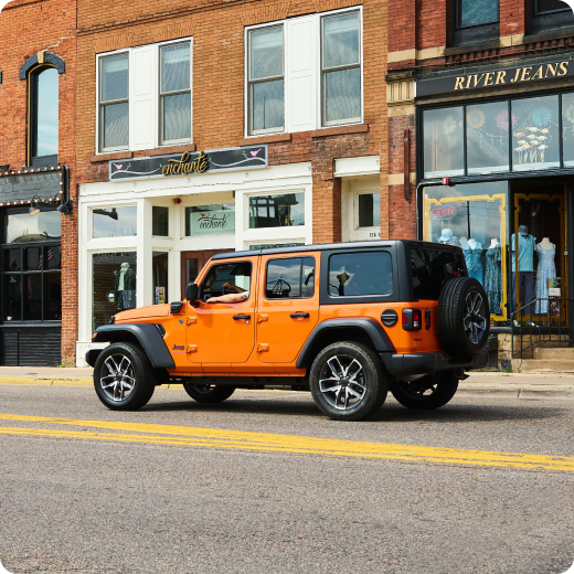 An orange SUV drives down a store-lined city street.
