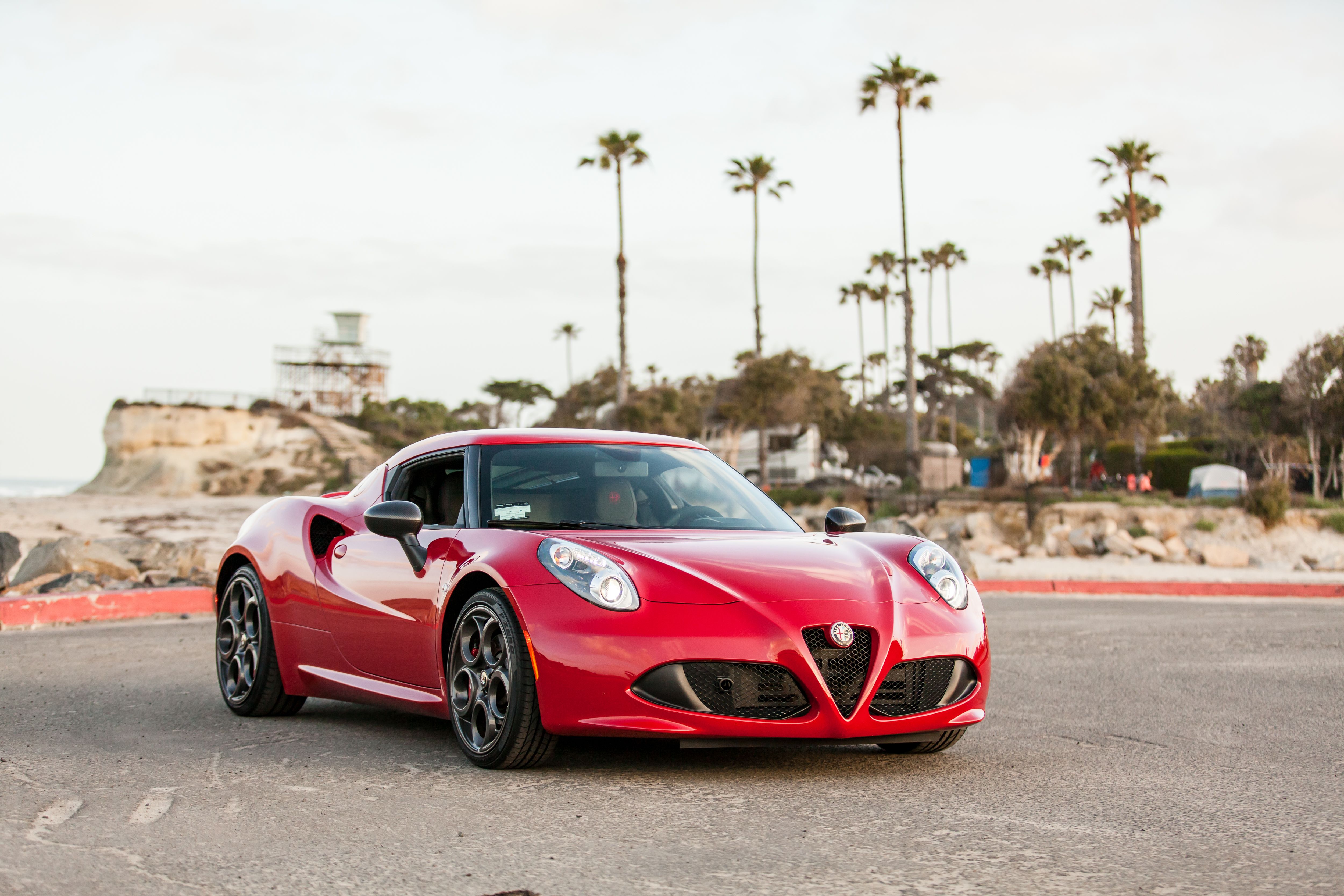 Red sports car parked by a palm-lined coastal road with a lifeguard tower in the background