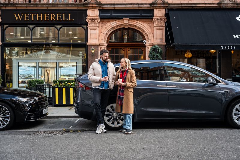 Couple with coffee beside a grey SUV on a Liverpool city street