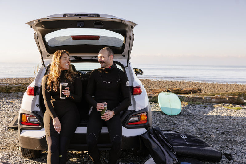 Two surfers in wetsuits sit in an open SUV trunk on a Cairns beach with surfboards nearby