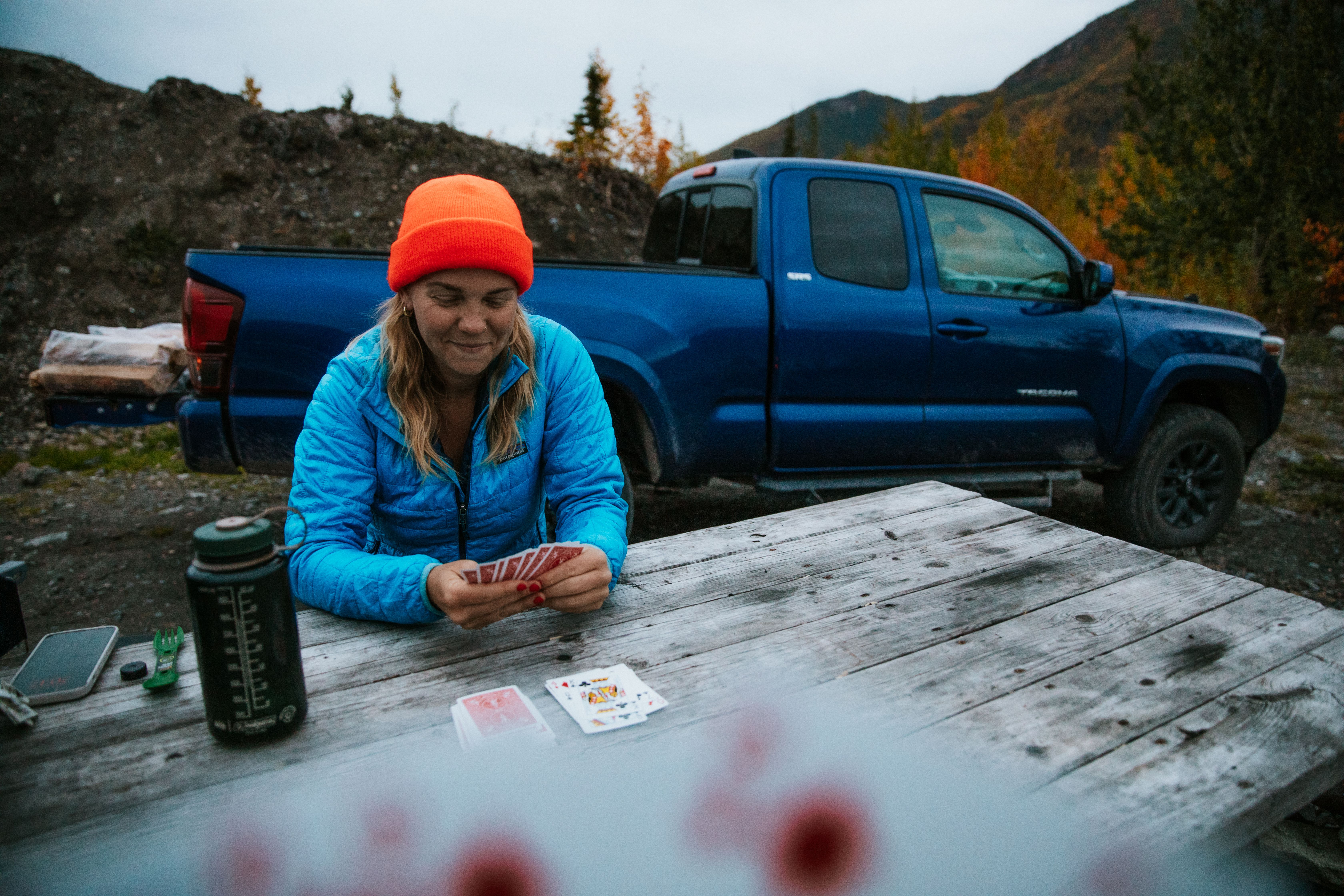 Woman in orange beanie plays cards at a picnic table beside a blue pickup in the Juneau mountains