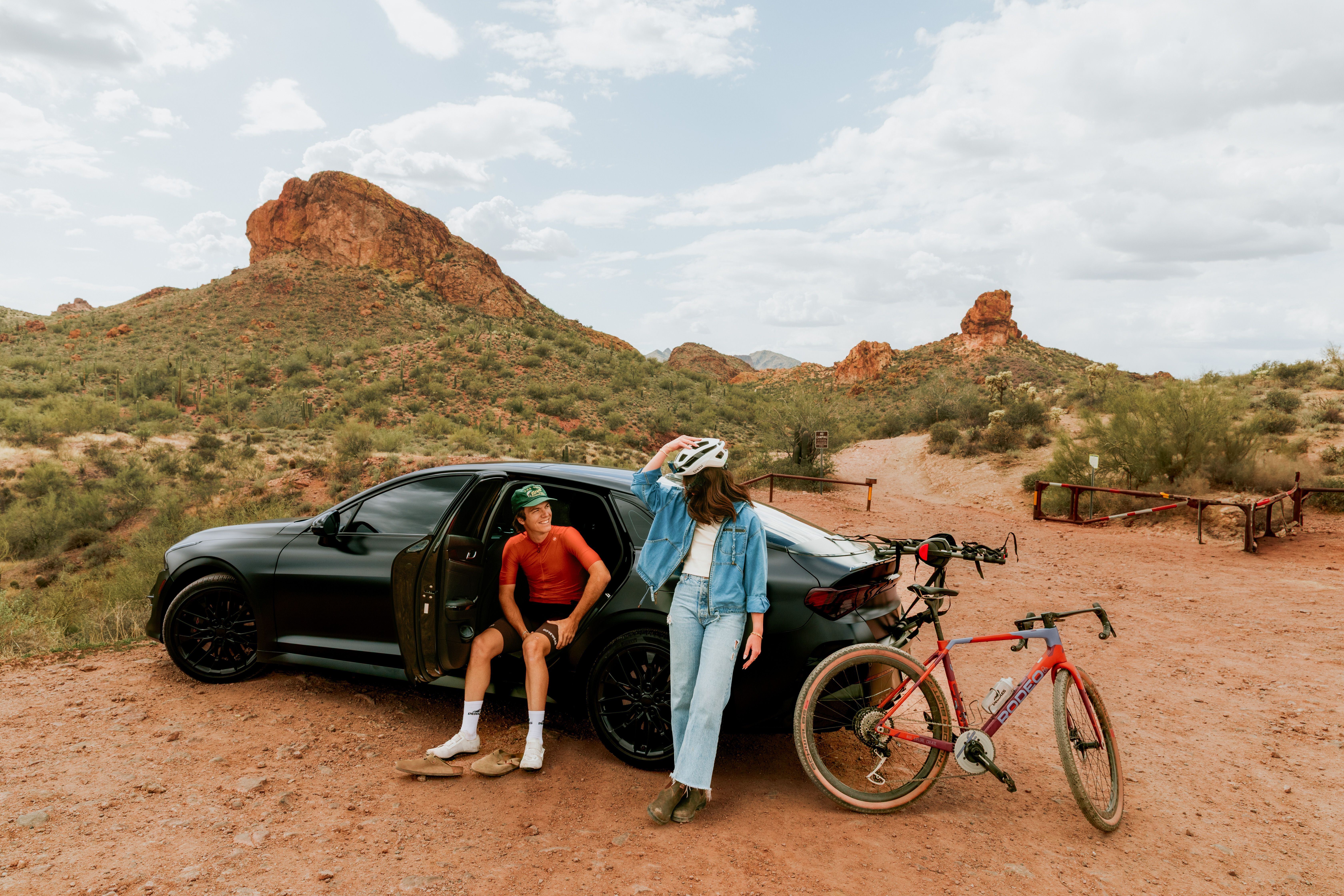 Cyclists by a black car in a red rock desert landscape with cacti and cloudy sky