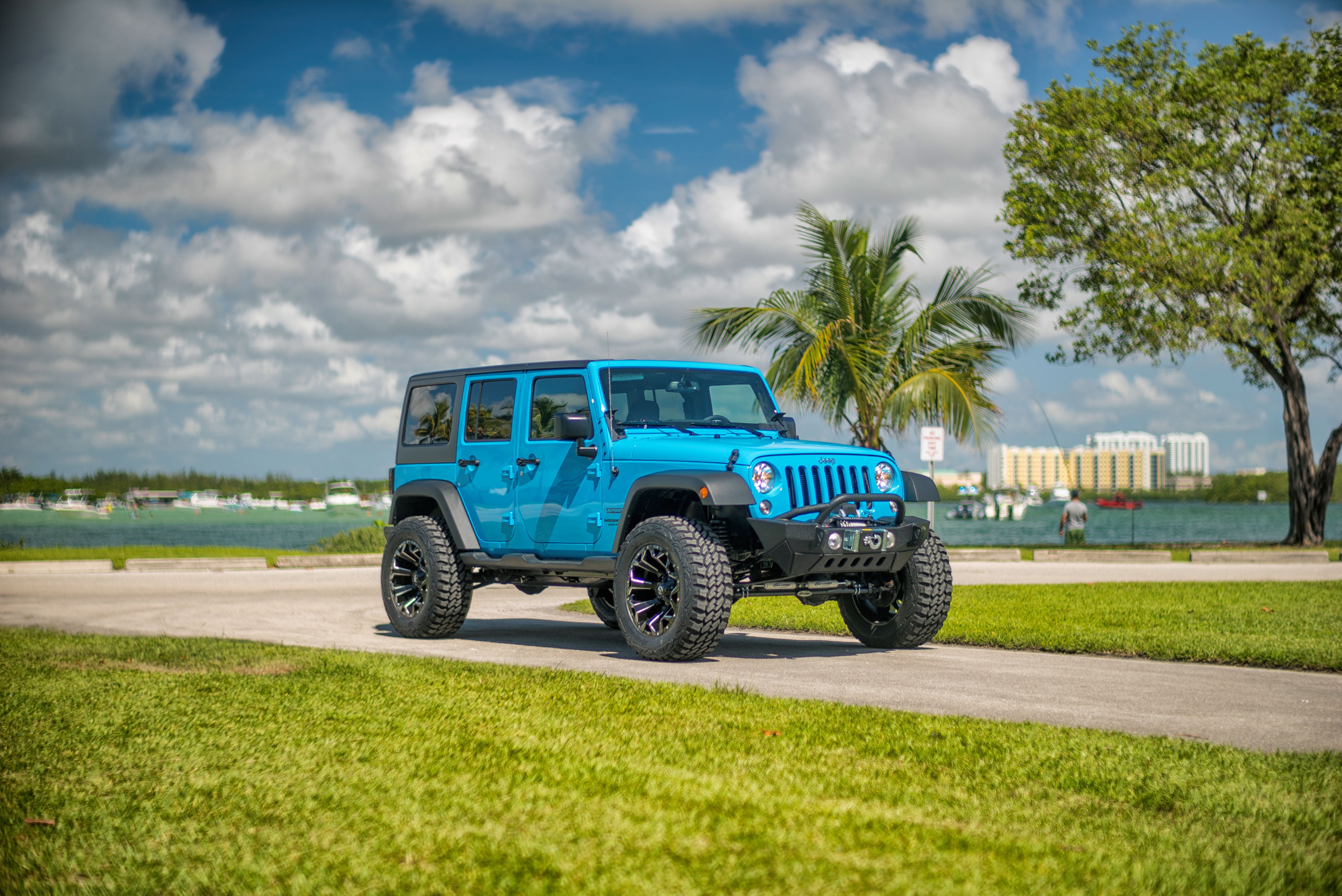 Blue Jeep parked by a Miami waterfront with palm trees, boats, and cloudy sky